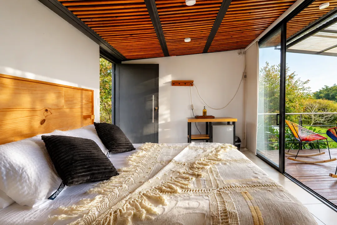 Cama King con cojines negros y vista a la terraza con sillas de colores en la cabaña