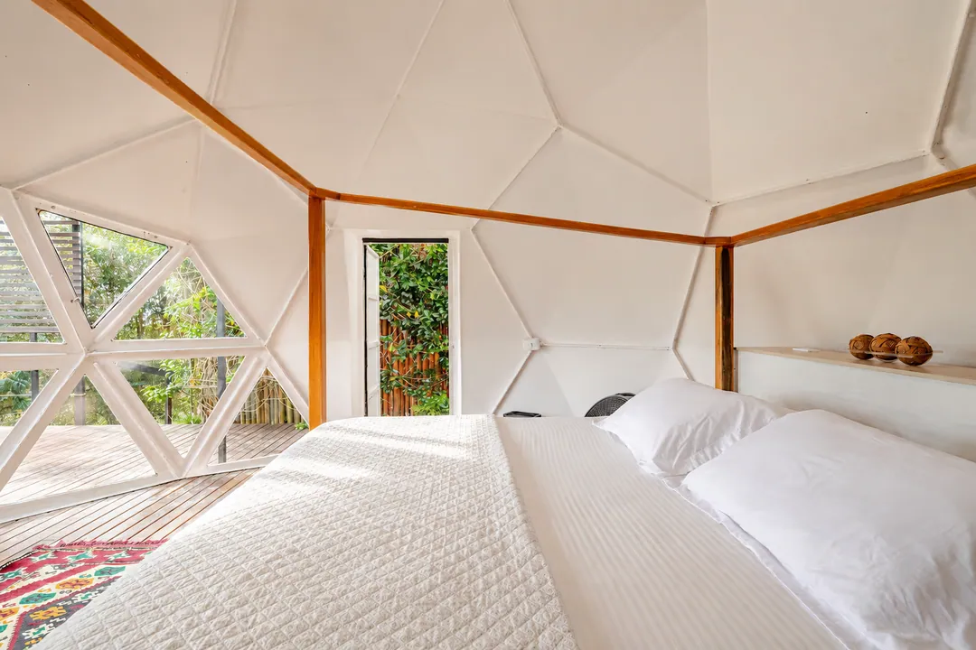 Cama King con ropa blanca dentro del Domo Ginger, con ventanales triangulares al jardín