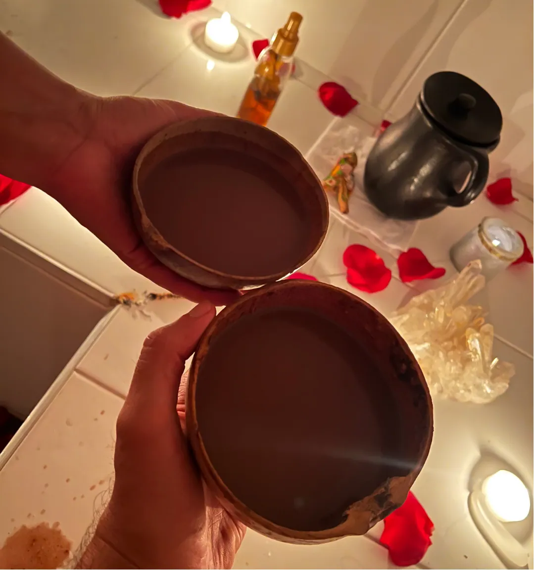 Dos manos sostienen tazas de barro con cacao, entre velas, pétalos de rosa y cristales