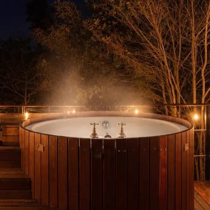 Jacuzzi de madera al aire libre con vapor, iluminado de noche entre árboles
